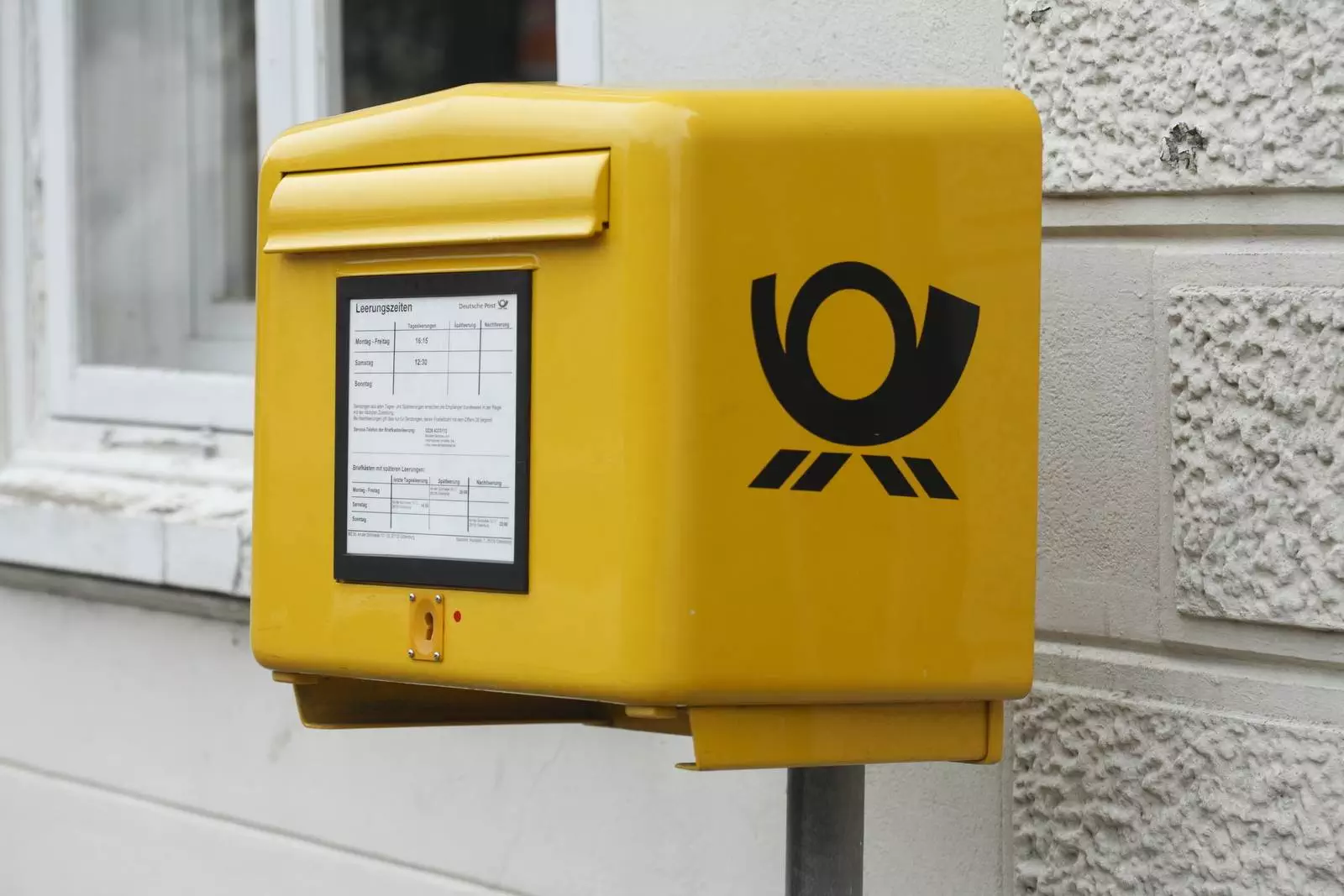 Ein leuchtend gelber Briefkasten mit schwarzem Postsymbol ist an einer Wand neben einem Fenster angebracht. Auf der Vorderseite des Briefkastens befindet sich eine Tafel mit Fahrplänen und Informationen.