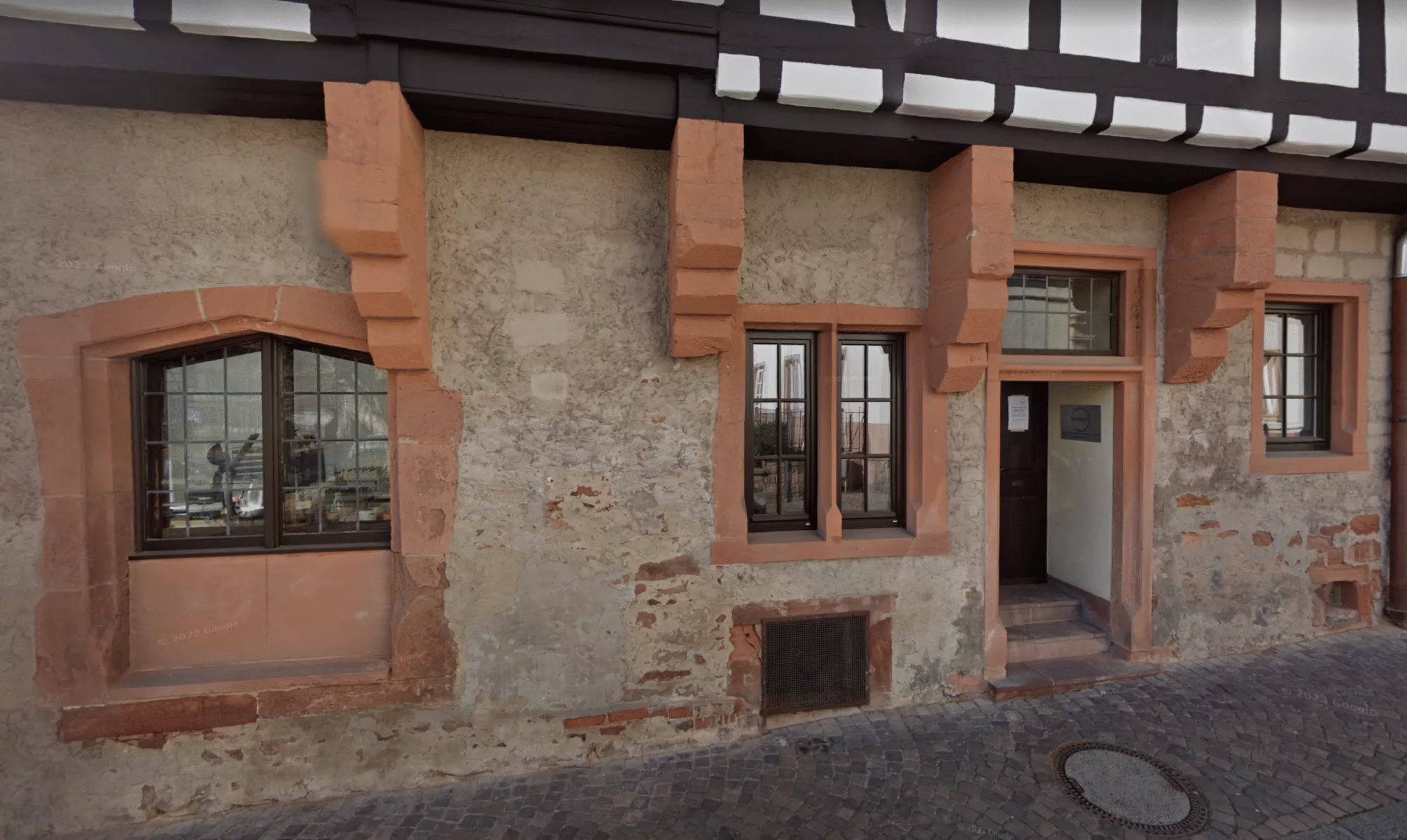 Ein rustikales Steingebäude mit braunen Fensterrahmen und einer Tür mit rötlichen Steinakzenten liegt an einer Kopfsteinpflasterstraße. Die Wand weist Witterungs- und Altersspuren auf.